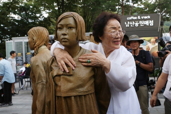 정면을 응시하며 손을 맞잡고 있는 세명의 소녀(한국, 중국, 필리핀)를 '위안부' 피해자 이용수 할머니가 끌어안았다.